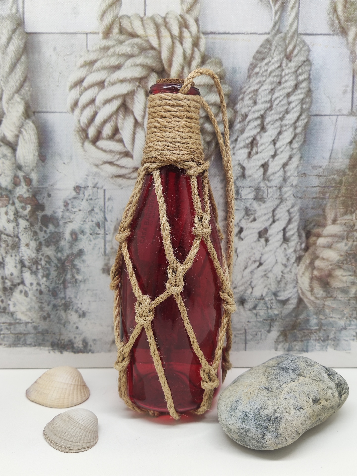 Garrafa de vidro vermelho envolvida em rede de corda natural ao lado de conchas e pedra com fundo decorativo.