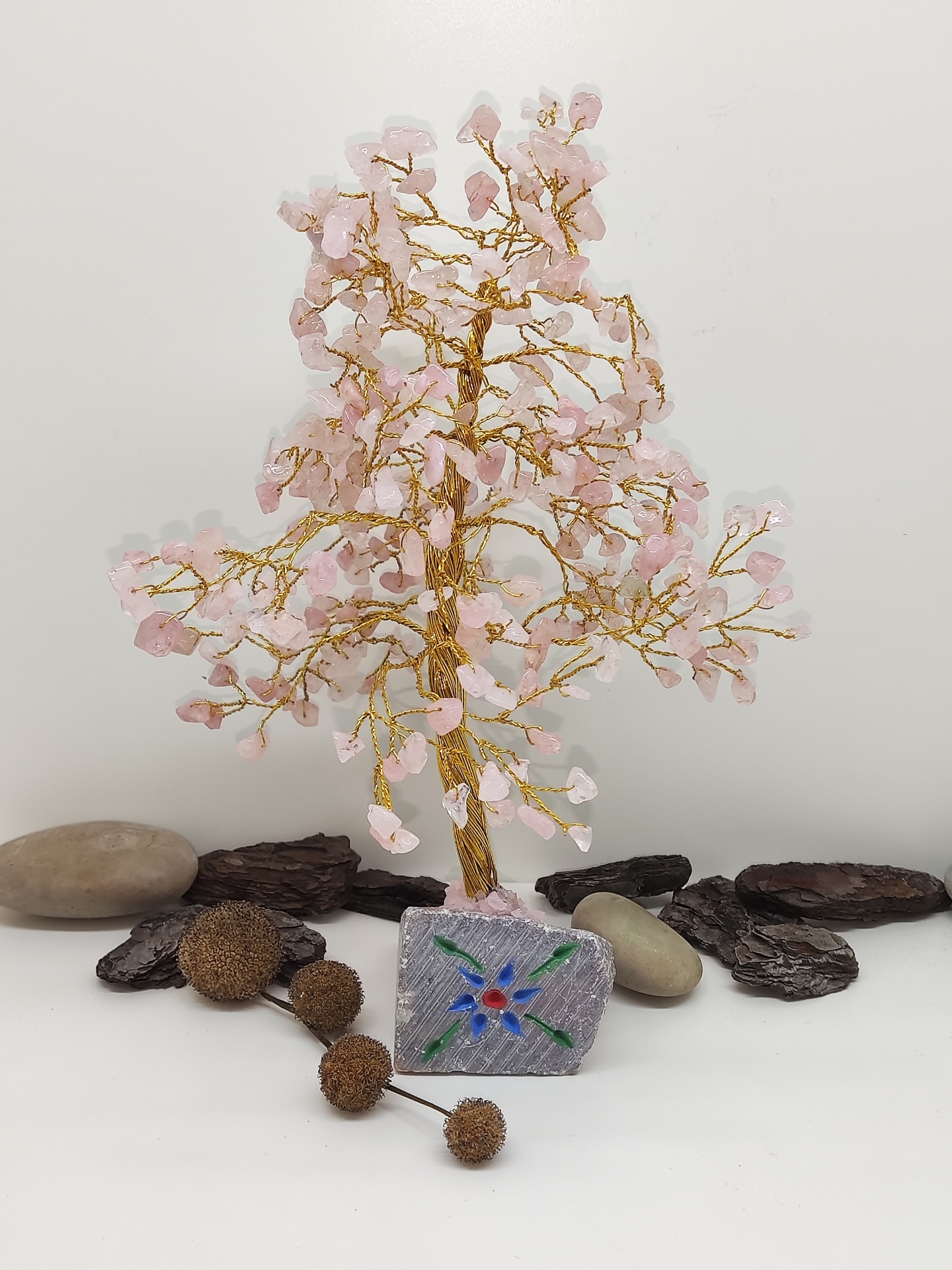 Árvore decorativa de fios dourados e pedras rosa com base quadrada decorada, em fundo branco com pedras e madeira