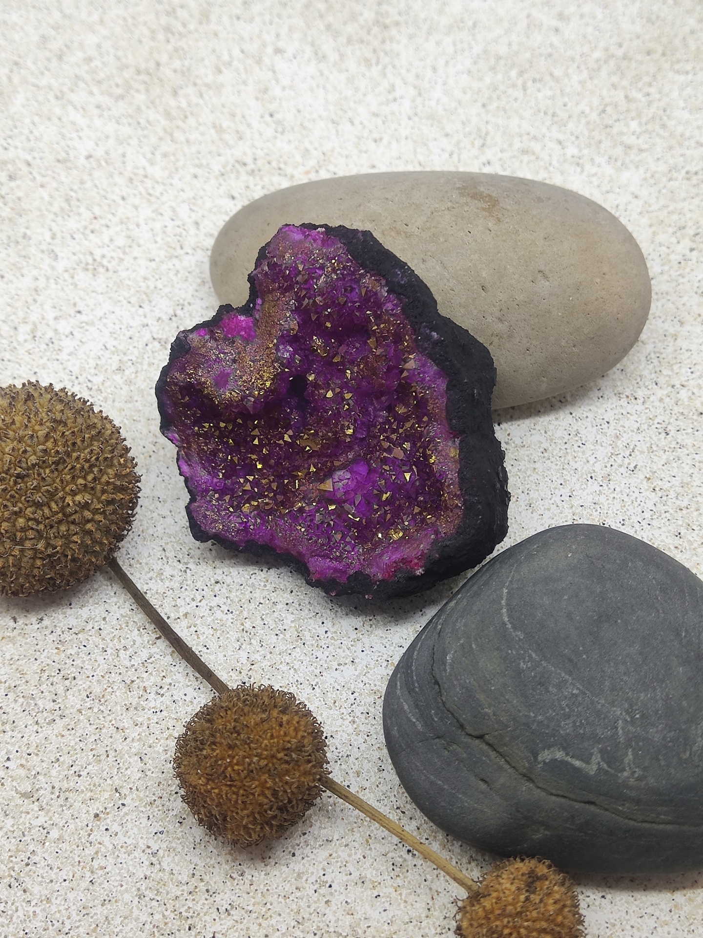 Geodo roxo com pedras lisas e planta seca sobre superfície clara.