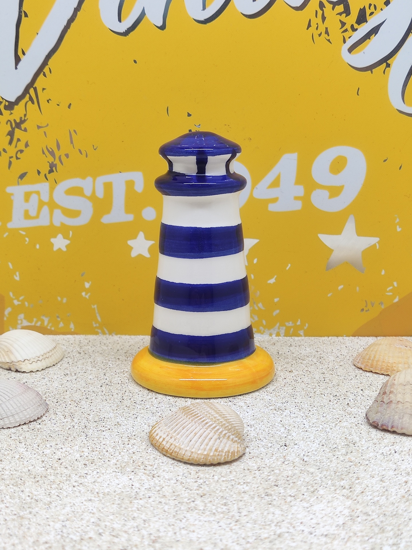 Farol decorativo azul e branco com base amarela e conchas sobre superfície granulada