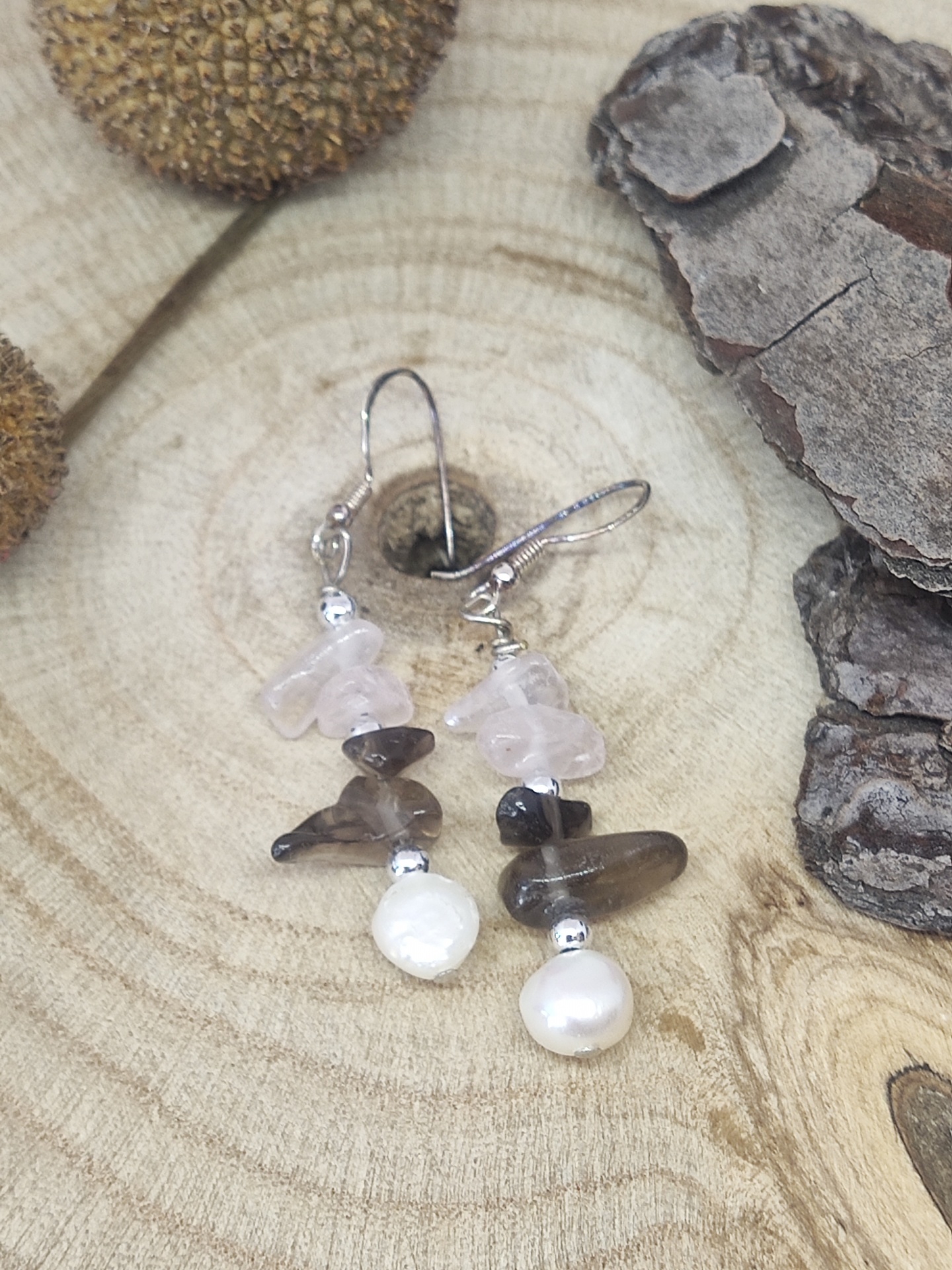 Brincos pendentes com pedras rosa e castanhas e pérola sobre madeira clara com casca e sementes