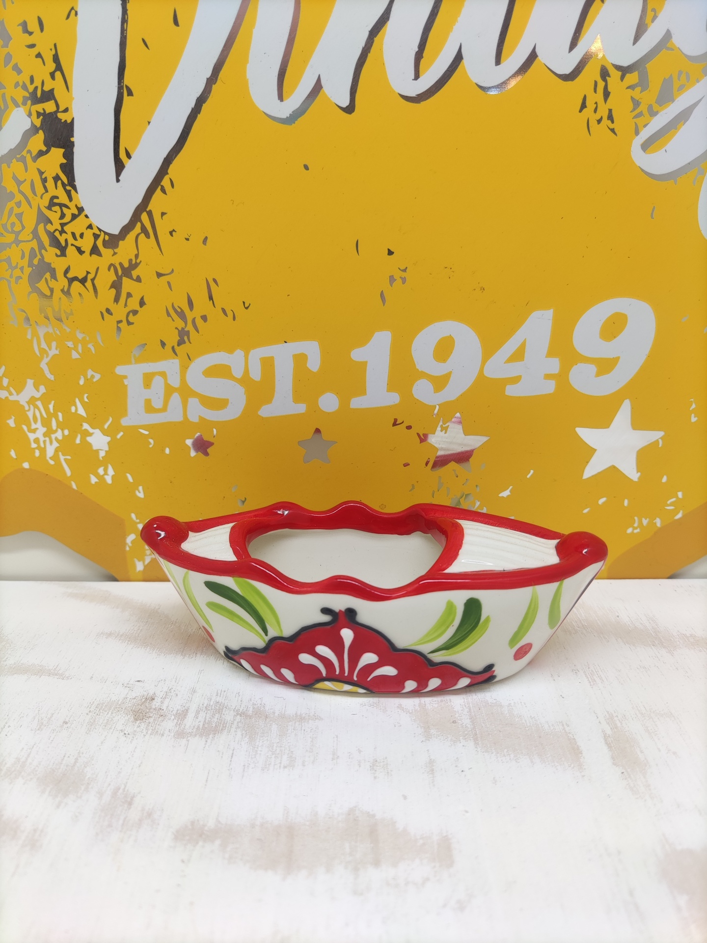 Cinzeiro cerâmica com padrão floral vermelho e verde na borda ondulada vermelha sobre fundo amarelo com texto EST. 1949