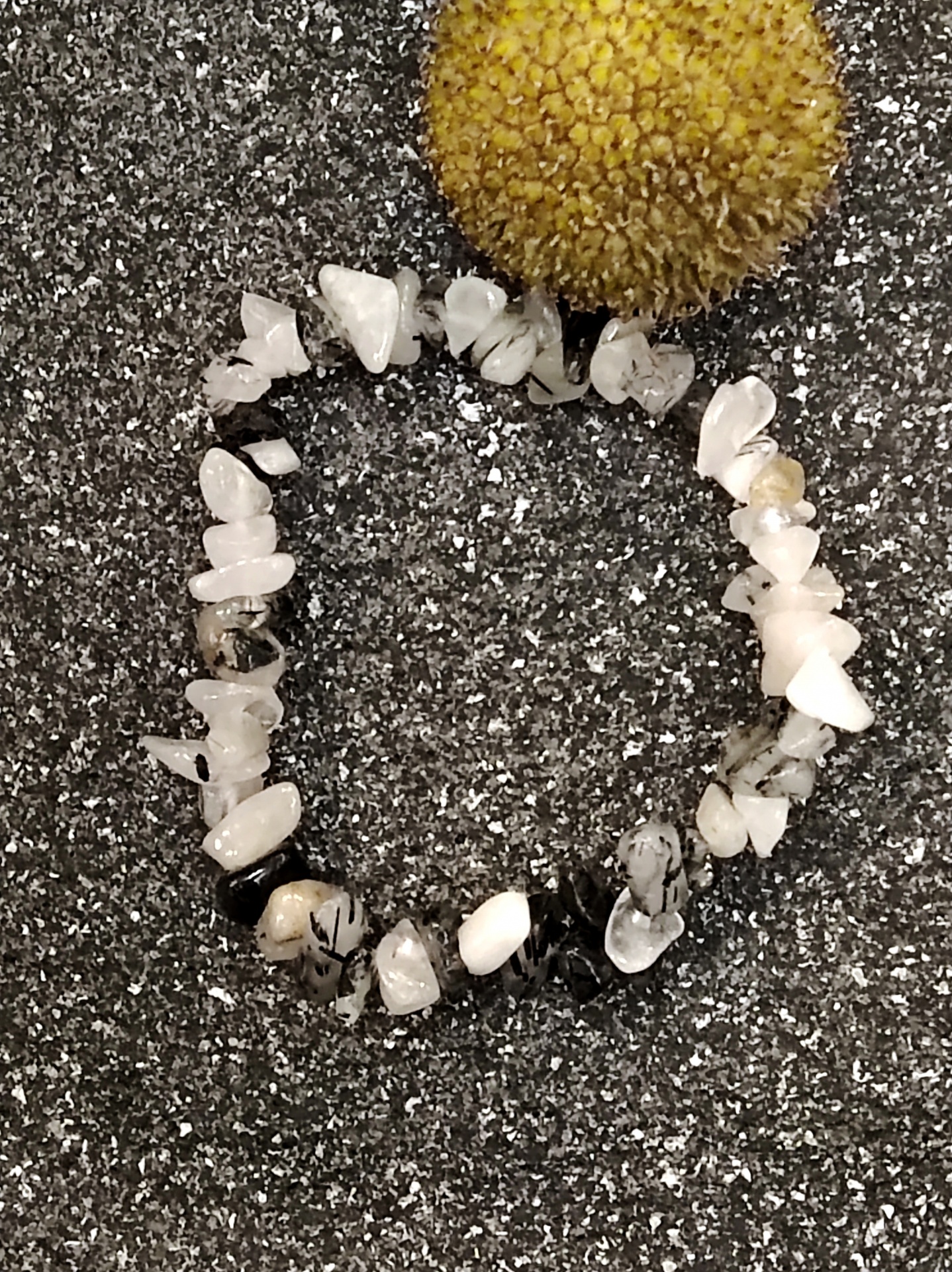 Pulseira de pedras brancas, pretas e bege sobre superfície cinzenta com flor amarela