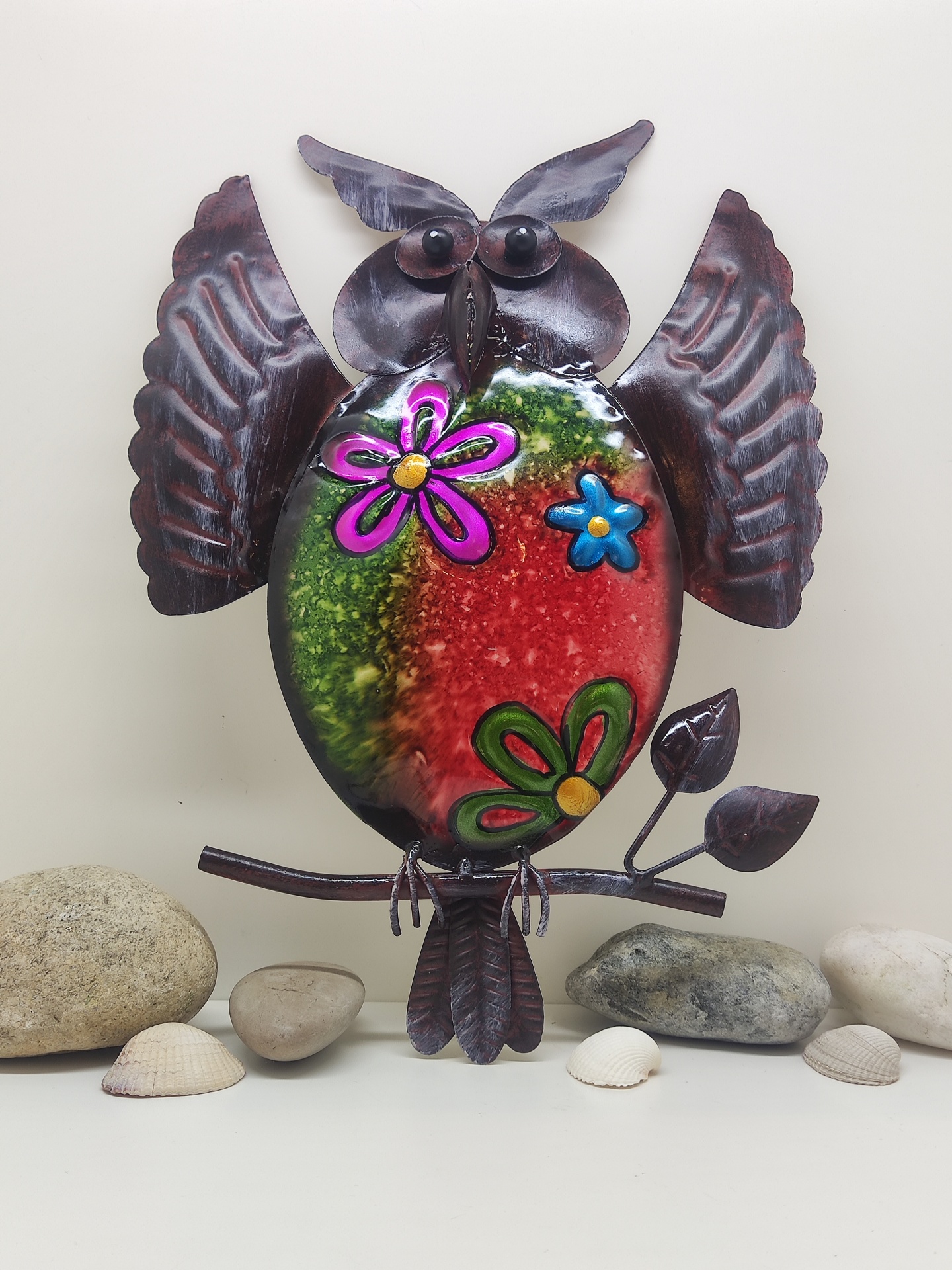 Escultura decorativa de coruja em metal colorido e castanho com flores sobre fundo com pedras e conchas.