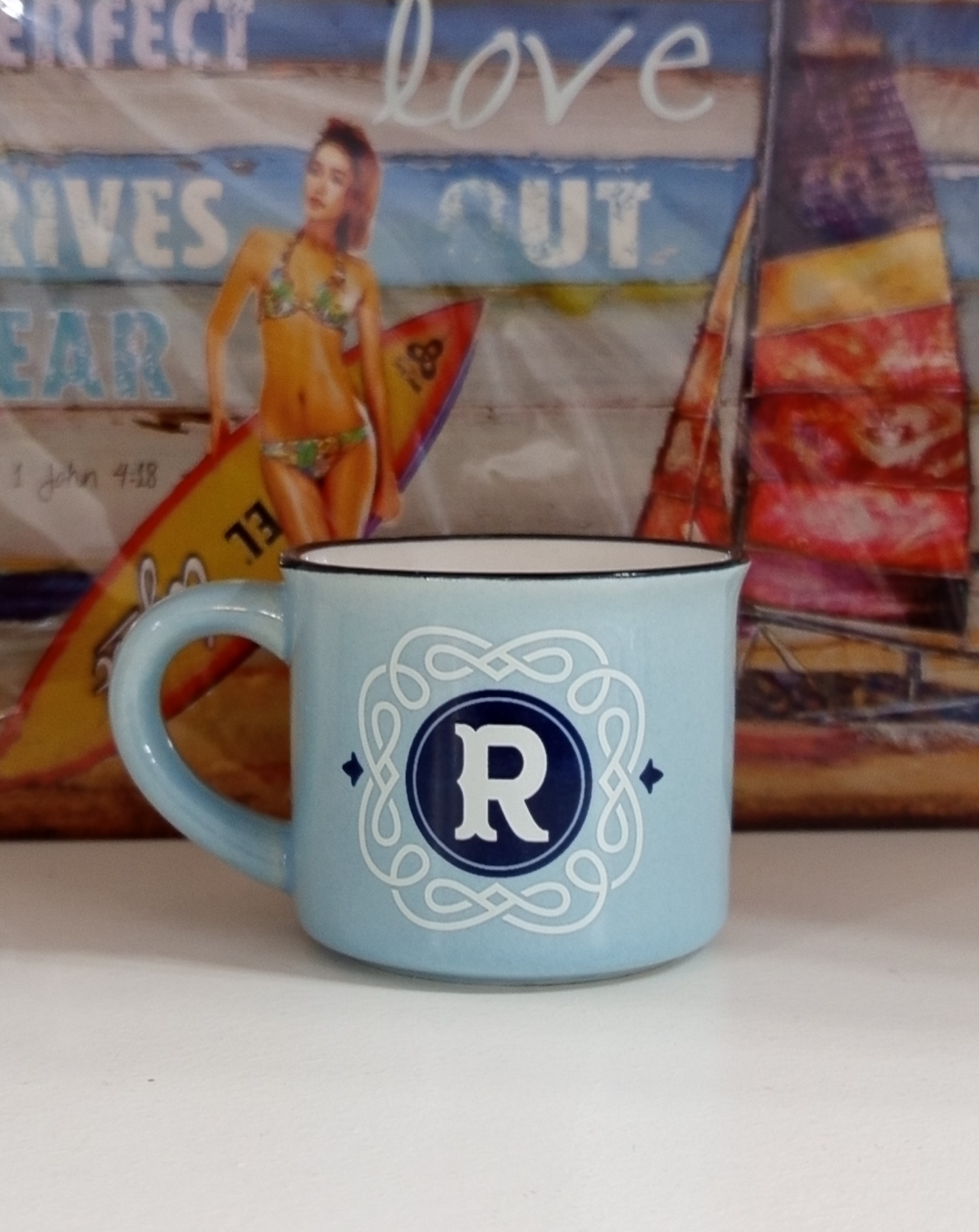 Caneca azul clara com letra 'R' e decoração branca em fundo de imagem com mulher de biquíni e prancha de surf