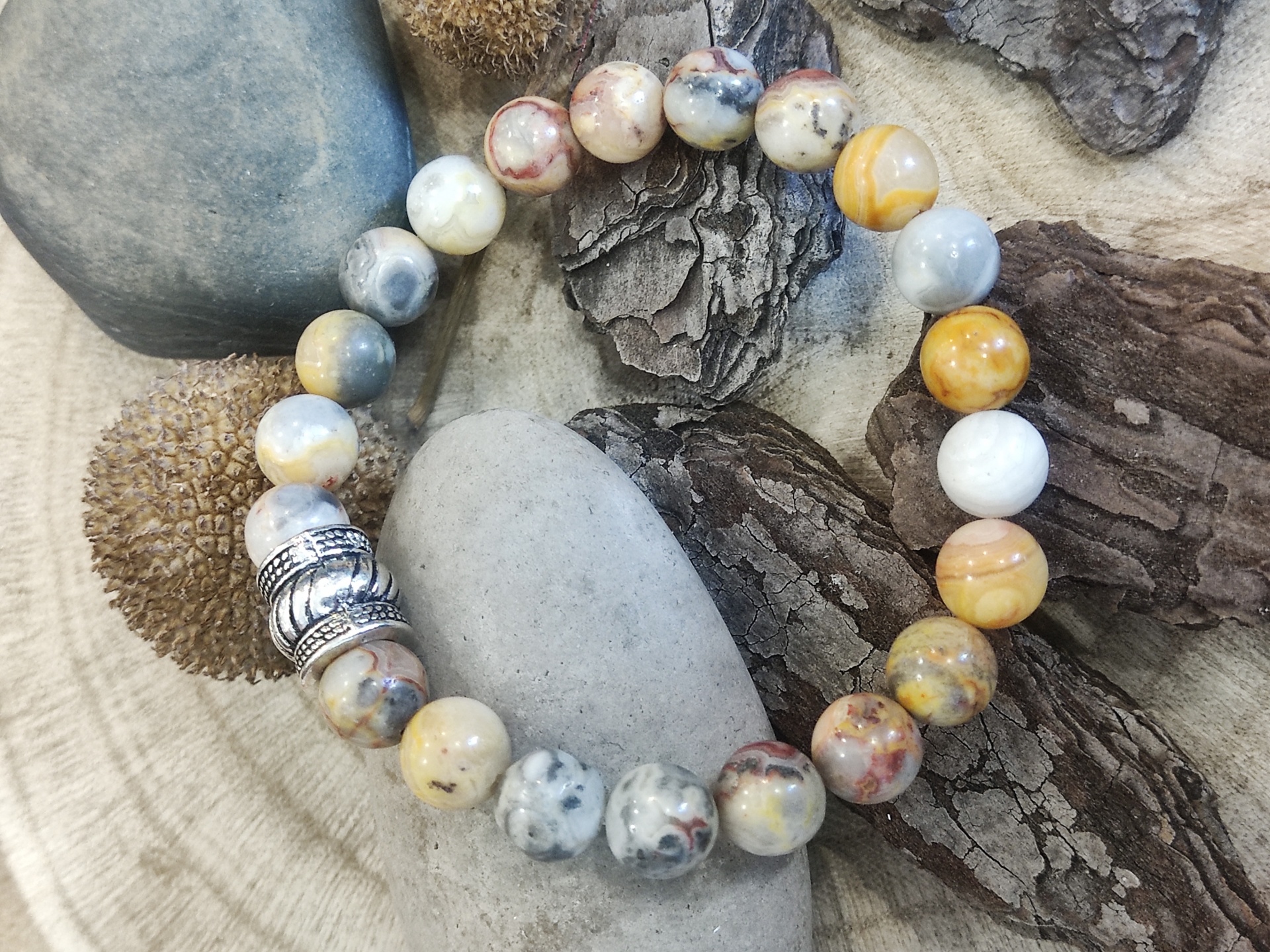 Pulseira de contas marmoreadas bege e cinza com elementos metálicos sobre pedras e casca de árvore