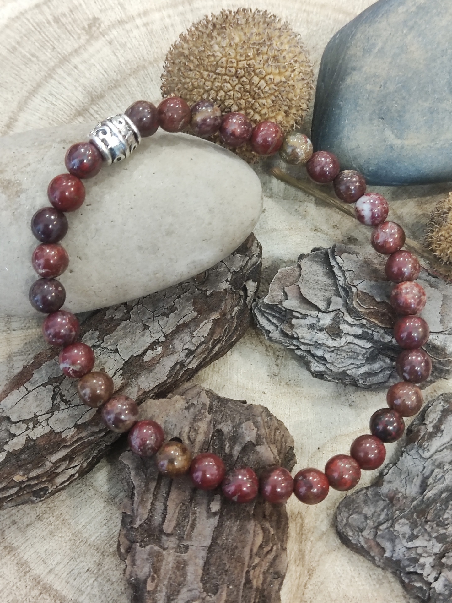 Pulseira de contas vermelhas, castanhas e bege com detalhe metálico, sobre casca de árvore e pedras