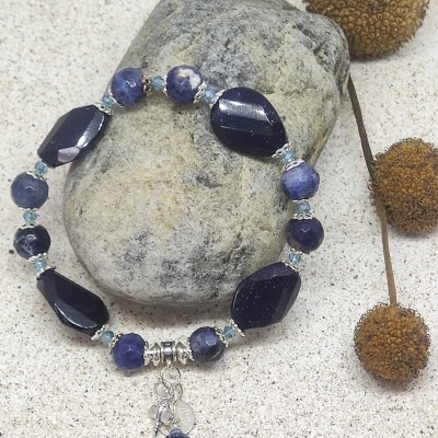 Pulseira azul com pingente sobre pedra cinzenta e fundo de areia clara com ramos secos