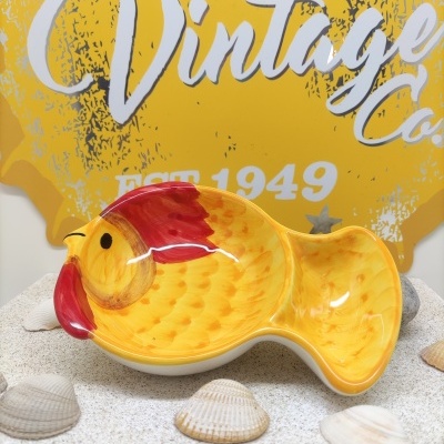 Prato em forma de peixe amarelo com detalhes vermelhos sobre areia com conchas e cartaz amarelo ao fundo