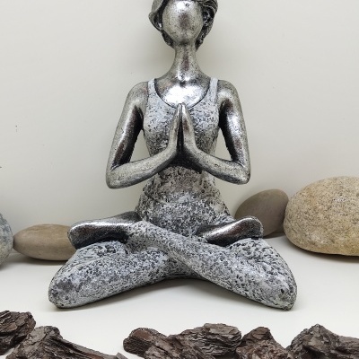 Estátua metálica prateada de mulher em posição de meditação com pedras e cascas ao redor