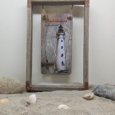 Quadro decorativo com farol e texto Algarve Portugal em madeira rústica pendurada dentro de moldura de madeira sobre areia com conchas e pedras