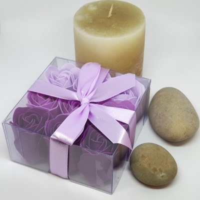 Caixa transparente com flores de sabonete lilás, vela creme e pedras ovais num fundo branco