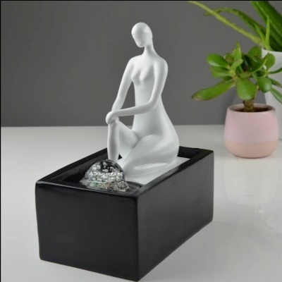 Fonte decorativa com figura feminina branca sobre base preta e planta suculenta ao fundo