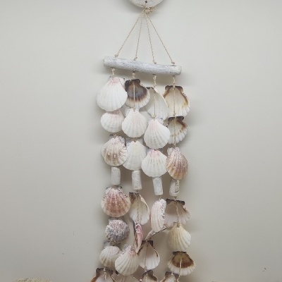 Móvel pendente feito de conchas naturais em tons claros e peças de madeira branca com detalhes azuis, pendurado numa parede branca, sobre areia e pedras.