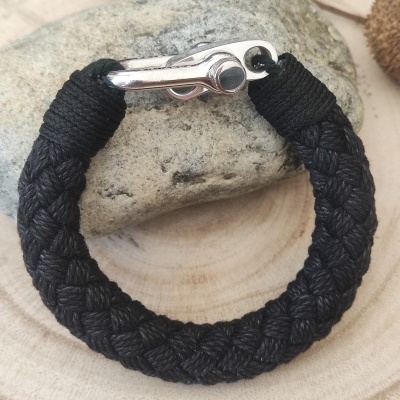 Pulseira de corda preta trançada com fecho metálico prateado sobre pedra e fundo de madeira