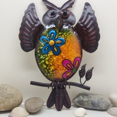 Estatueta de coruja decorativa em metal com padrão colorido e flores, rodeada por pedras e conchas.