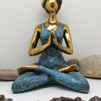 Estátua decorativa dourada e azul de mulher em pose de meditação com pedras e madeira ao redor