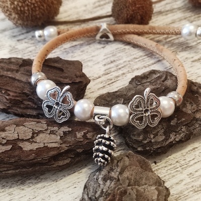 Pulseira em cortiça com contas brancas, ornamentos florais metálicos e pendente de pinha sobre fundo de madeira e casca