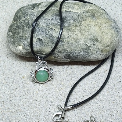 Colar com pendente de caranguejo prateado e pedra verde em cordão preto sobre pedra e areia