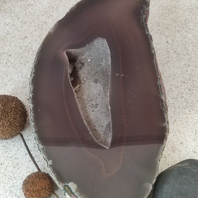 Peça decorativa de ágata castanha com cavidade, ao lado de pedras e plantas secas sobre superfície branca