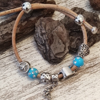 Pulseira em cortiça com contas metálicas prateadas e azuis, com pendente de menina, sobre madeira clara e cascas