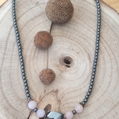 Colar com contas metálicas prateadas, contas rosa claro e conta preta oblonga sobre tábua de madeira clara