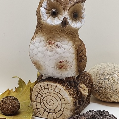 Estatueta de coruja castanha e branca sentada num tronco decorativo com elementos naturais ao redor