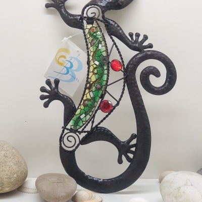 Escultura metálica de lagarto com mosaico verde e vermelho junto a pedras e conchas