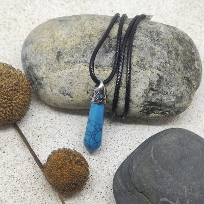 Colar com pendente de pedra azul turquesa e cordão preto sobre fundo de areia clara com pedras e elementos secos.