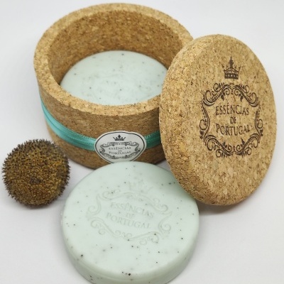 Sabonete branco redondo com partículas, caixa de cortiça redonda com etiqueta e tampa gravada, e fruta seca marrom ao lado