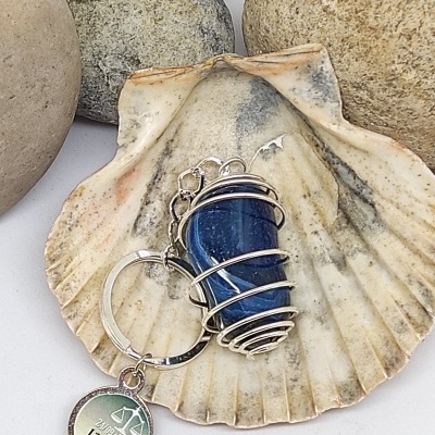 Chaveiro com pedra azul e pendente redondo sobre concha com pedras no fundo.