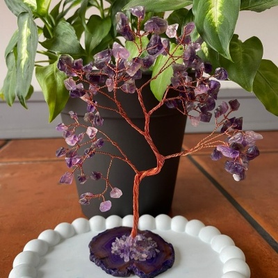 Árvore de fio de cobre com pedras roxas numa base branca, com planta verde em vaso preto ao fundo