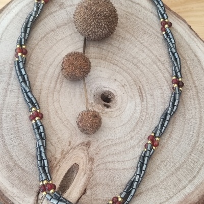 Colar com contas metálicas prateadas e esferas vermelhas e douradas sobre base de madeira clara