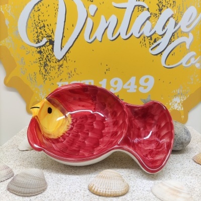 Prato cerâmico em forma de peixe vermelho com detalhes amarelos sobre areia e conchas, fundo amarelo com texto 'Vintage Co. EST. 1949'