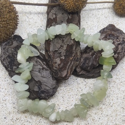 Pulseira de pedras verdes irregulares sobre areia com casca de árvore castanha e plantas secas.