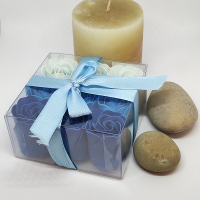 Vela castanha clara, pedras naturais e caixa transparente com flores de sabão azuis e brancas amarrada com fita azul