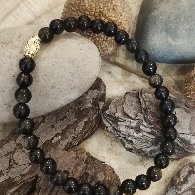 Pulseira de contas pretas com detalhe dourado sobre troncos, pedras e frutos secos