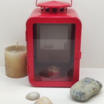 Lanterna metálica vermelha com vela castanha, conchas e pedra sobre fundo branco