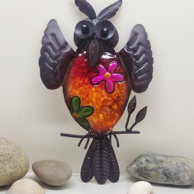 Escultura colorida de coruja em metal decorada com flores, sobre superfície com pedras e conchas.