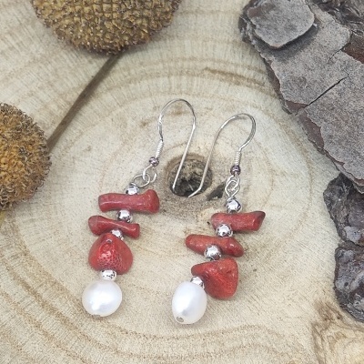 Brincos com pedras vermelhas, contas prateadas e pérolas brancas sobre madeira e elementos naturais.