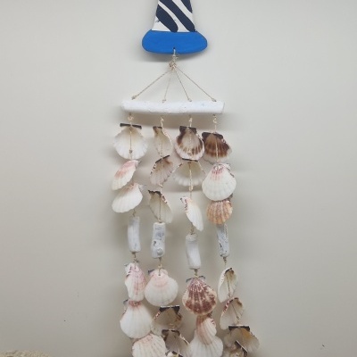 Suspensão decorativa com conchas, madeira pintada de azul e branco, pendurada numa parede clara sobre areia e pedras