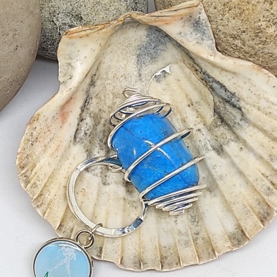 Chaveiro com pedra azul e pendente 'Acuario' numa concha creme com pedras ao fundo.