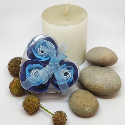 Conjunto decorativo com vela branca, flores de tecido azuis e pedras cinzentas sobre fundo branco.