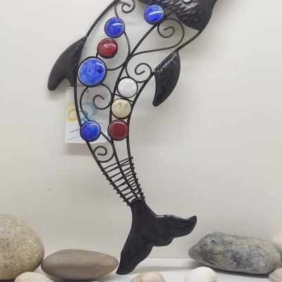 Escultura de golfinho em metal com detalhes coloridos colocada sobre uma superfície branca com pedras e conchas.