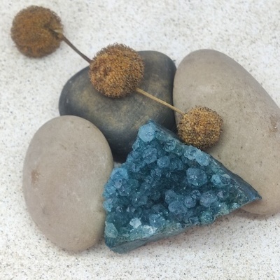 Pedra mineral azul com cristais sobre pedras lisas e flores secas em superfície arenosa.