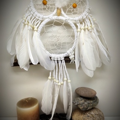 Pendurado decorativo em forma de coruja com penas brancas e miçangas amarelas, vela e pedras ao lado.
