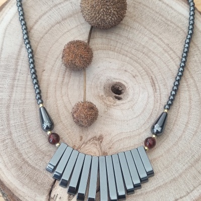 Colar em contas escuras com pendentes cinza metálico sobre base de madeira clara com frutos secos.