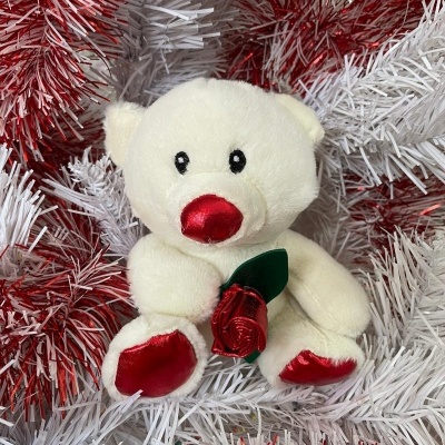 Ursinho de peluche branco com nariz e patas vermelhas segurando rosa vermelha sobre decoração de ramos brancos e vermelhos.