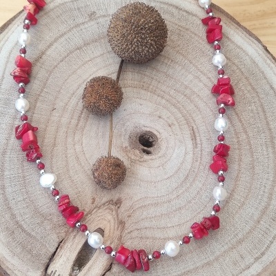 Colar com pedras vermelhas, pérolas e contas prateadas sobre madeira clara com bolas secas.