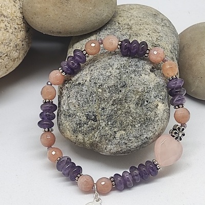 Pulseira de pedras em tons castanho-avermelhado e roxo com pendente metálico, sobre pedras lisas cinzentas